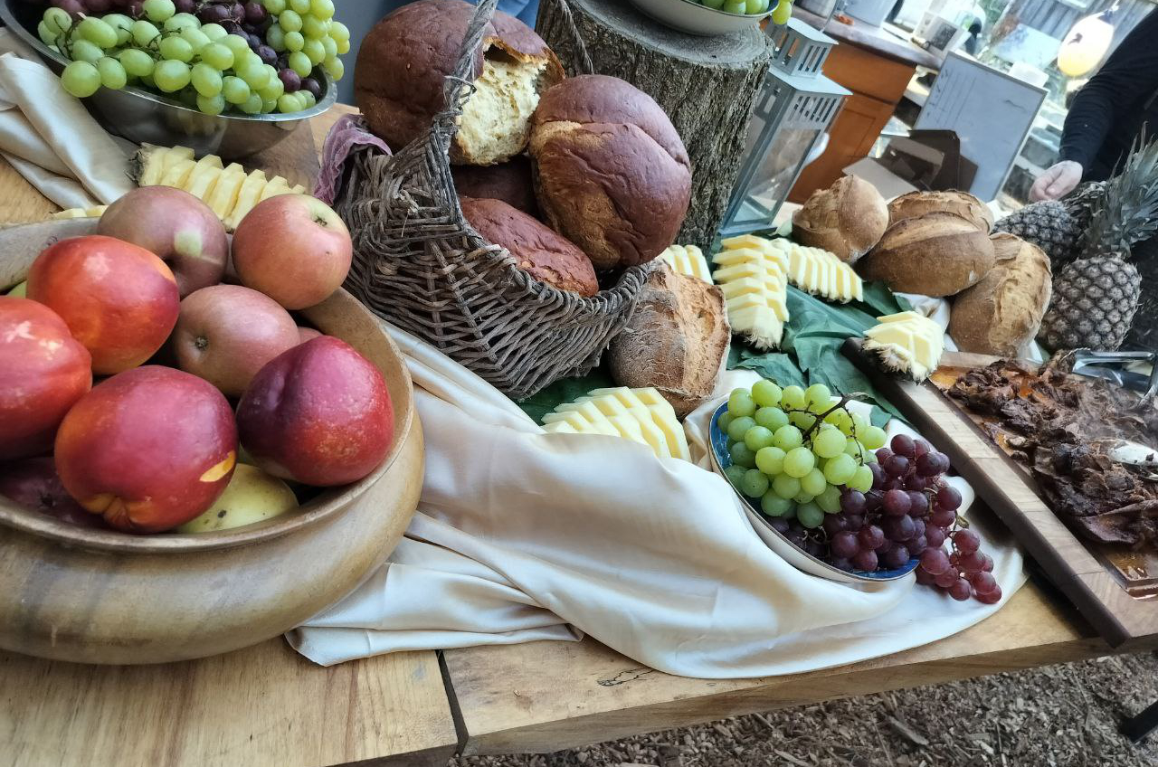 A feast of food laid out on a wooden table, including apples in a wooden bowl, a basket of bread, sliced and whole pineapples, and roasted meat on a wooden cutting board, surrounded by an ecru scarf.