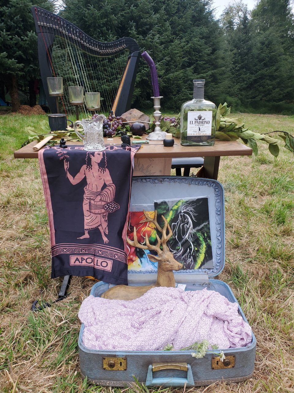An altar laid out on a wooden table. Atop the table is a glass ewer holding up a black-and-earthen brown banner of Apollo; a purple candle; leaves and grapes. Behind the altar stands a large black harp. In front, an open suitcase bears colorful art and a statue of a seated stag.