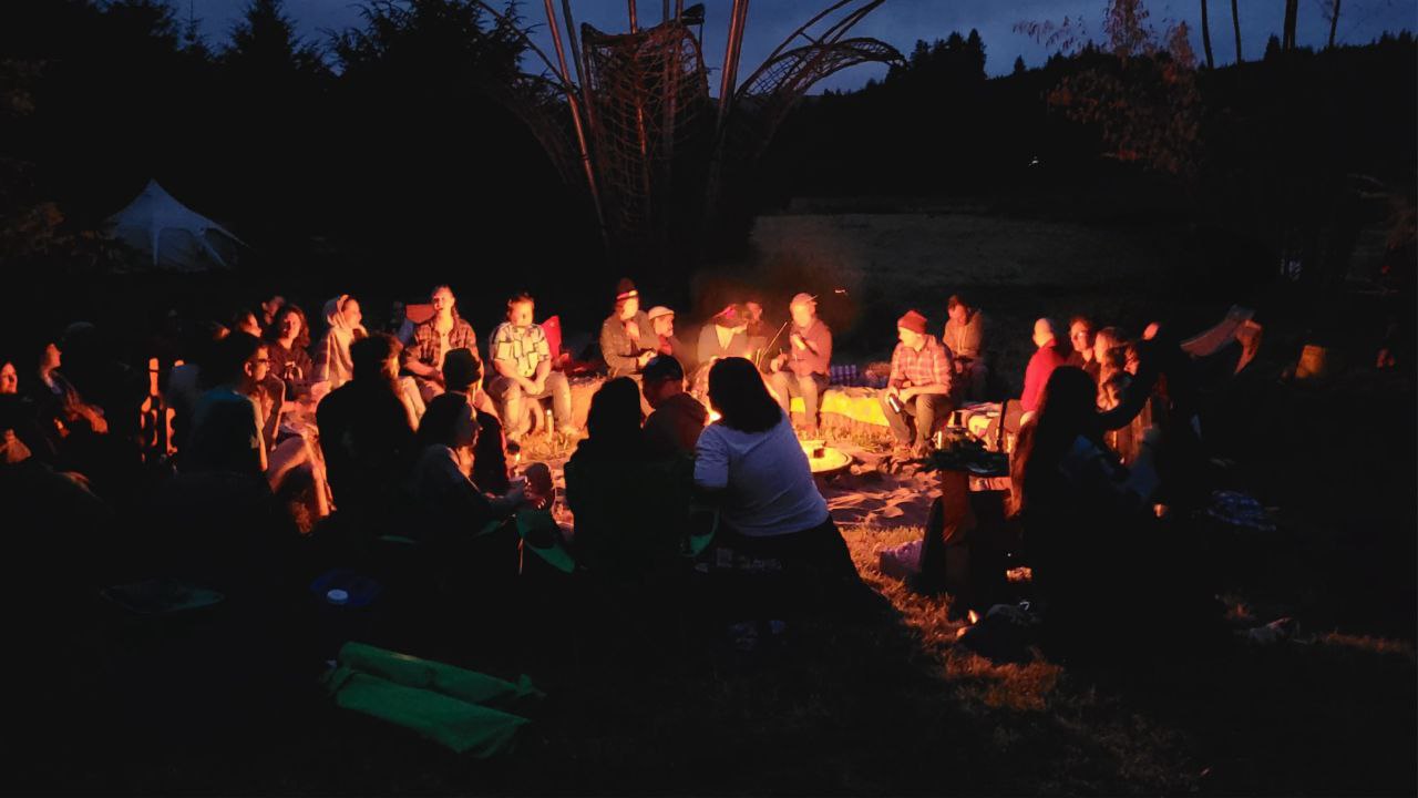 A group of people, slightly blurred by motion, sit in a circle around a fire in the dark.