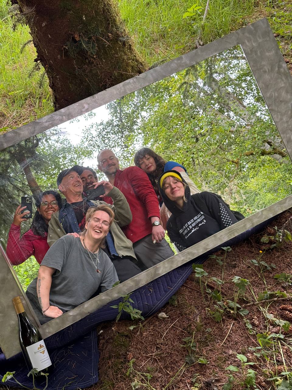 A group of friends takes a selfie in a mirror propped up on a tree, a bottle of wine in the foreground.